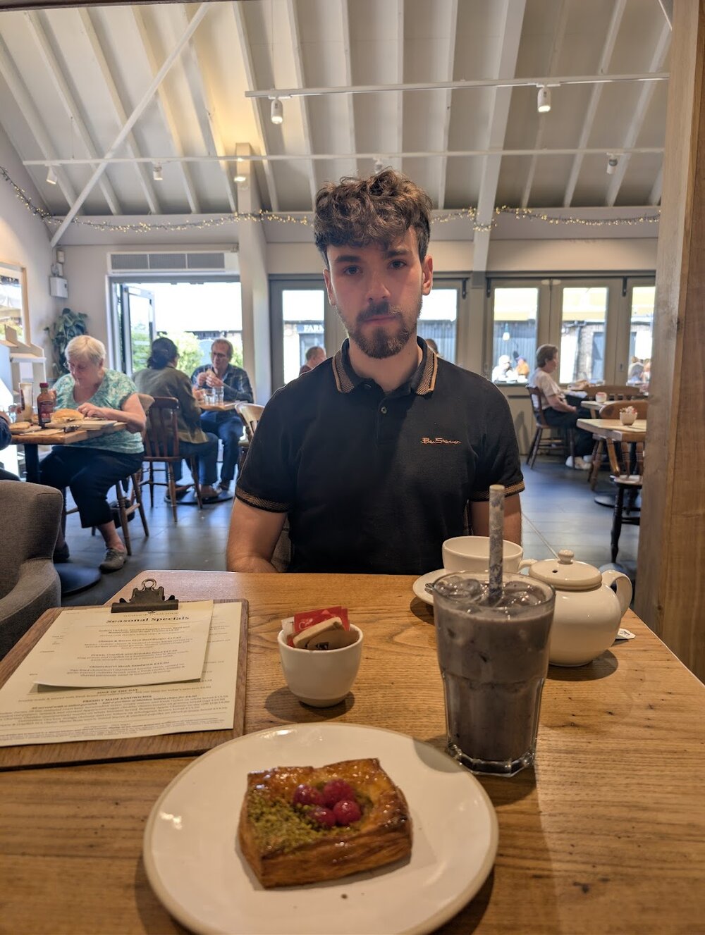 Jason Harding sitting expressionless at a café table with a pastry and drinks